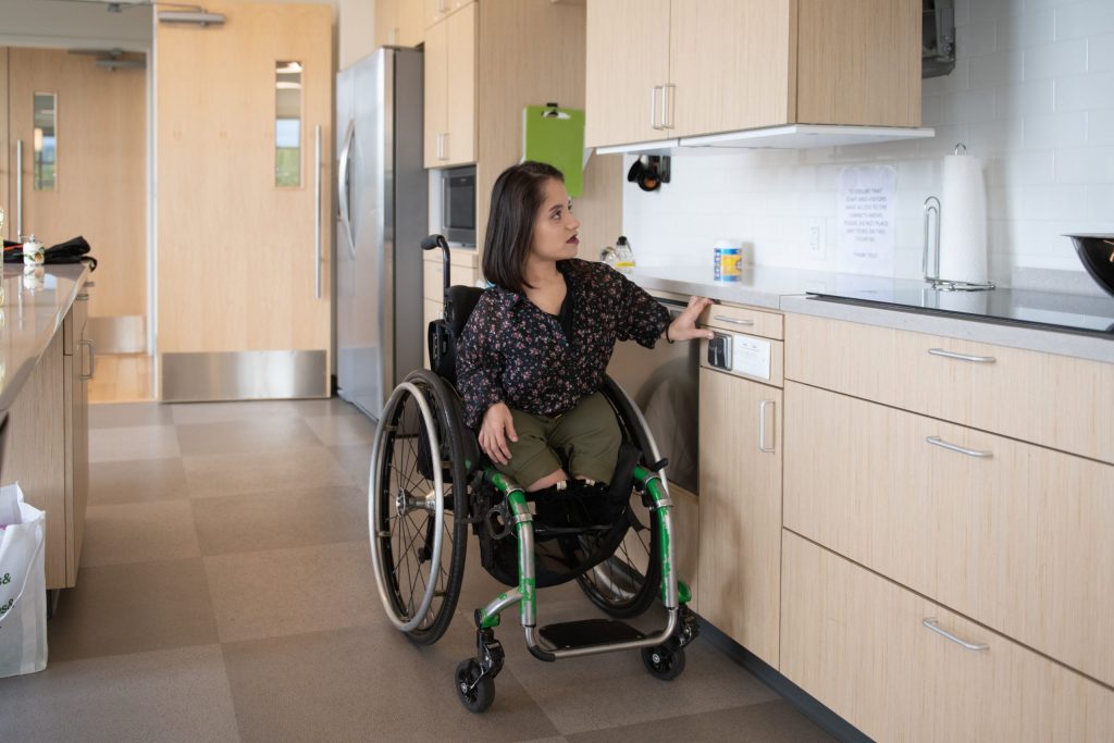 A South Asian person sits in her wheelchair and presses a button to lower the height-adjustable shelves in an accessible kitchen.