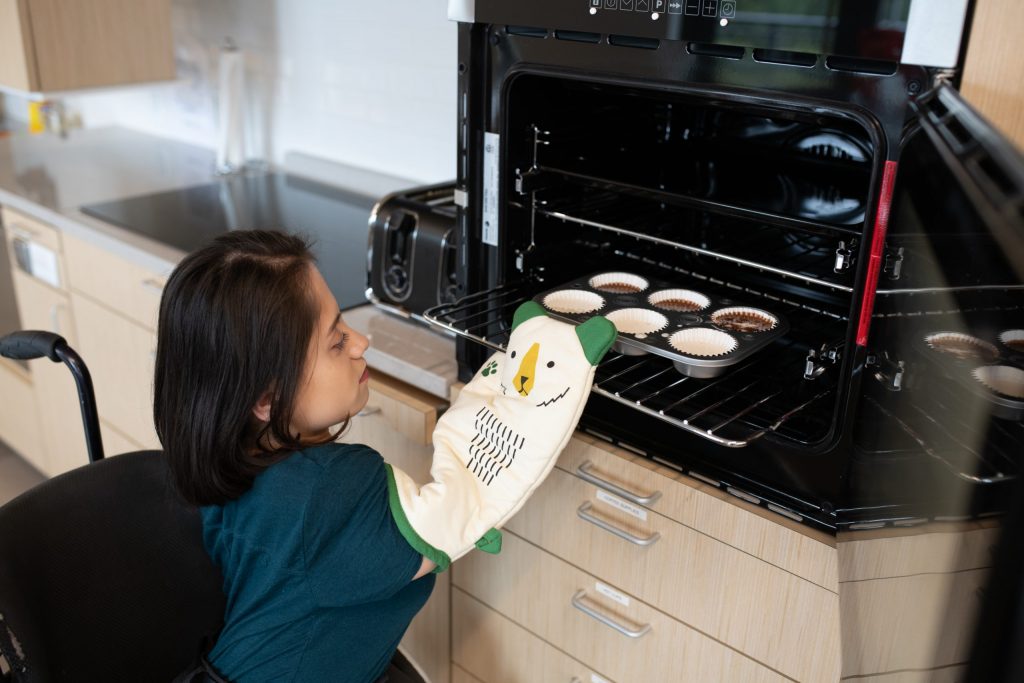 A South Asian person sits in her wheelchair and places a muffin pan into a mid-height oven with side-hinged door. She is wearing an oven mitt designed to look like a bear waving its paw.