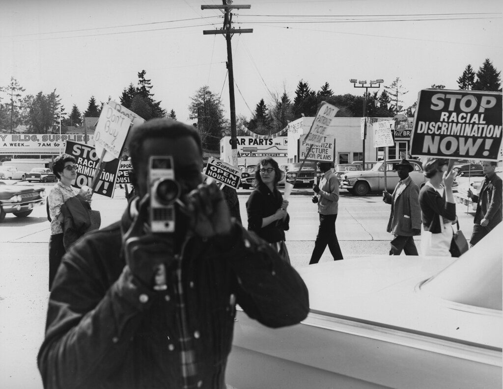 Photo of a man holding a camera at a fair housing protest in 1964. Behind him protesters hold signs with phrases like, 